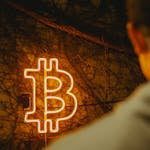 Glowing bitcoin symbol in neon light on vine wall with person in foreground, suggesting finance themes.